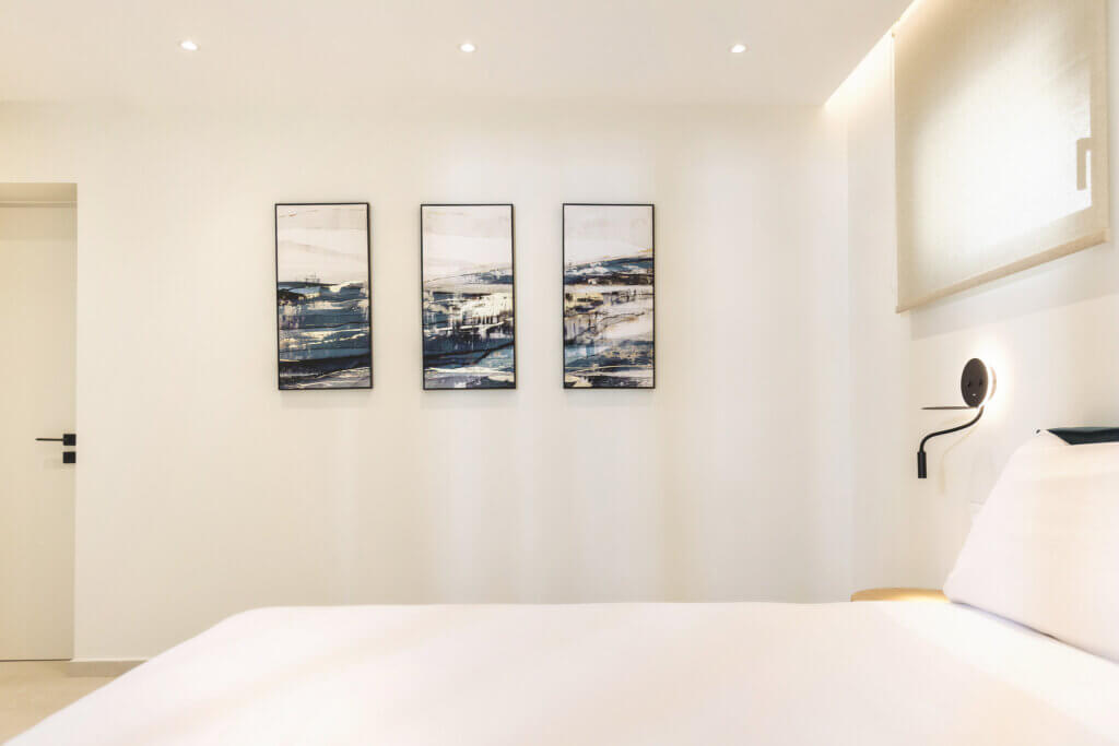 This image showcases a section of a bedroom featuring a comfortable bed in the foreground, with a set of three framed abstract paintings hanging above it, bringing artistic flair to the space. The warm lighting from the bedside lamp casts a soft glow, enhancing the room’s welcoming ambience.