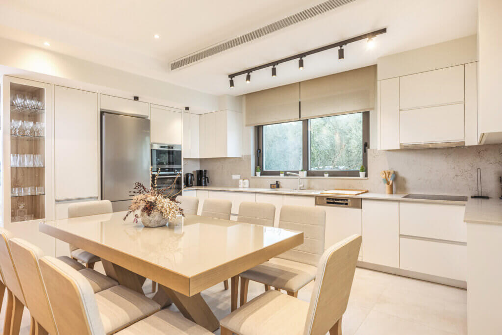 An open-plan kitchen with state-of-the-art appliances and a white dining table set for six under spotlights.