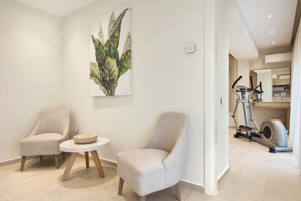 Two gray armchairs and a round table in a bedroom corner with a tropical leaf painting, adjacent to a workout area with fitness equipment.