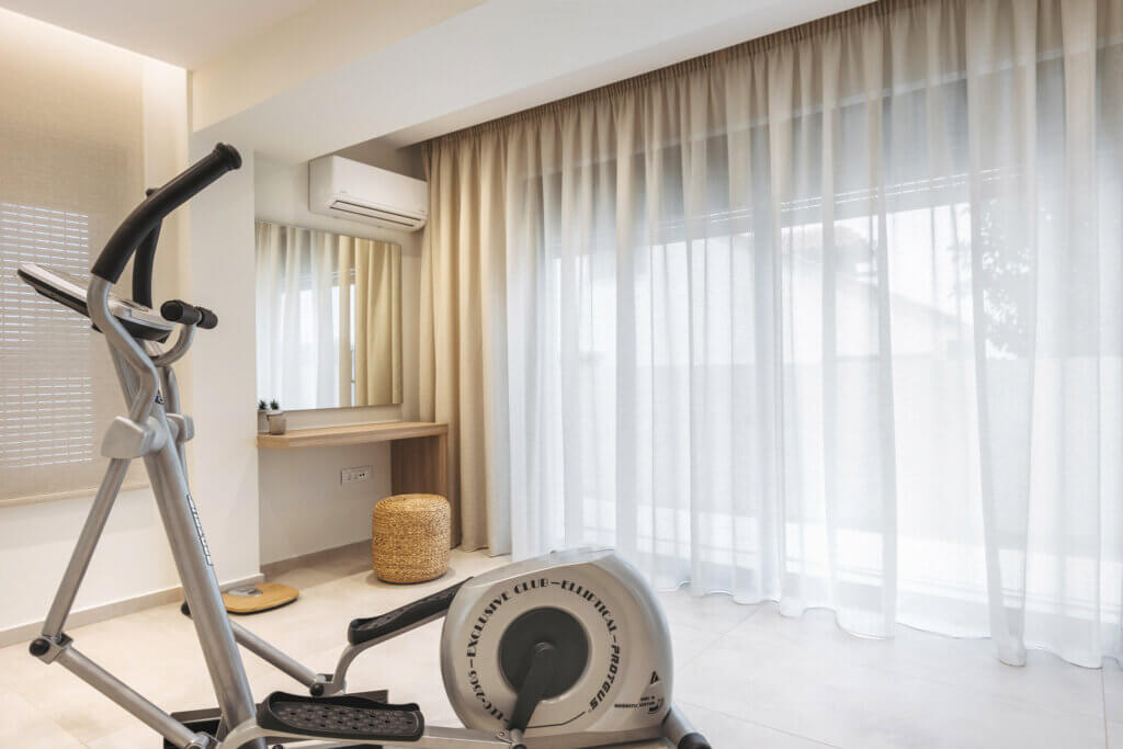 Exercise equipment in a bright villa bedroom with floor-to-ceiling windows covered by light curtains.